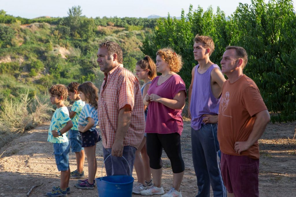 Grupo de personas de diferentes edades reunidas en un huerto, con árboles y tierra de cultivo al fondo