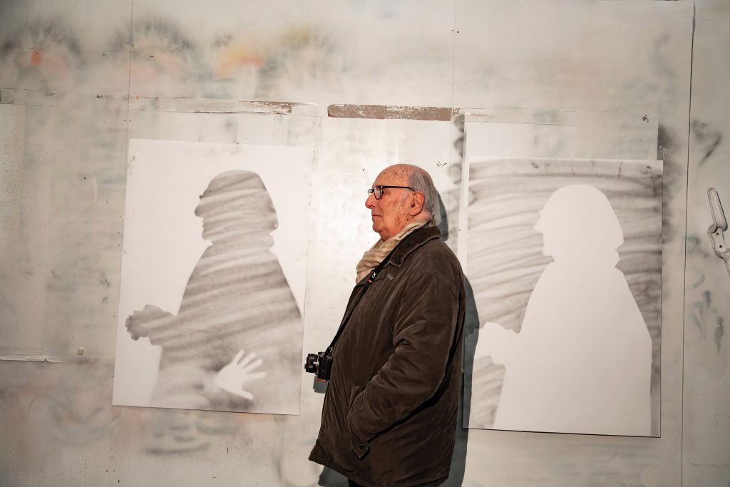 Carlos Saura observa dibujos de rostros en blanco y negro expuestos en una pared de taller