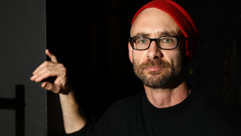 Chuck Palahniuk sosteniendo un micrófono durante una presentación, vistiendo gafas y gorro rojo