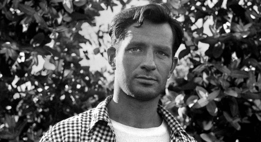 Retrato en blanco y negro de Jack Kerouac entre hojas de árbol, vistiendo camisa cuadriculada