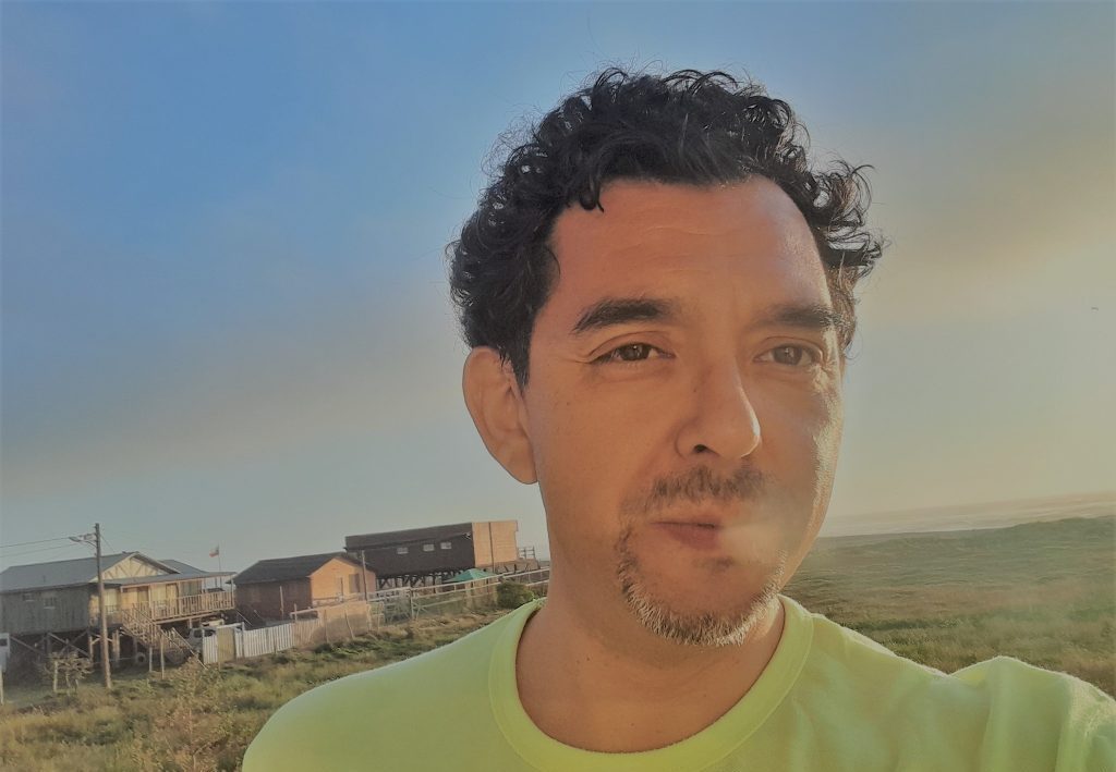 Hombre de mediana edad en playera verde claro frente a paisaje rural con construcciones al fondo