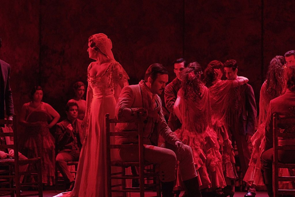 Escena teatral en rojo con actriz en primer plano vistiendo vestido largo, rodeada de actores en atmósfera dramática
