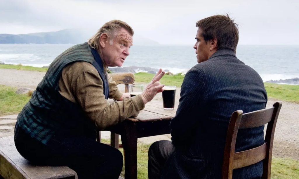 Dos hombres conversan alrededor de una mesa con bebidas, con vistas al mar y paisaje costero de fondo