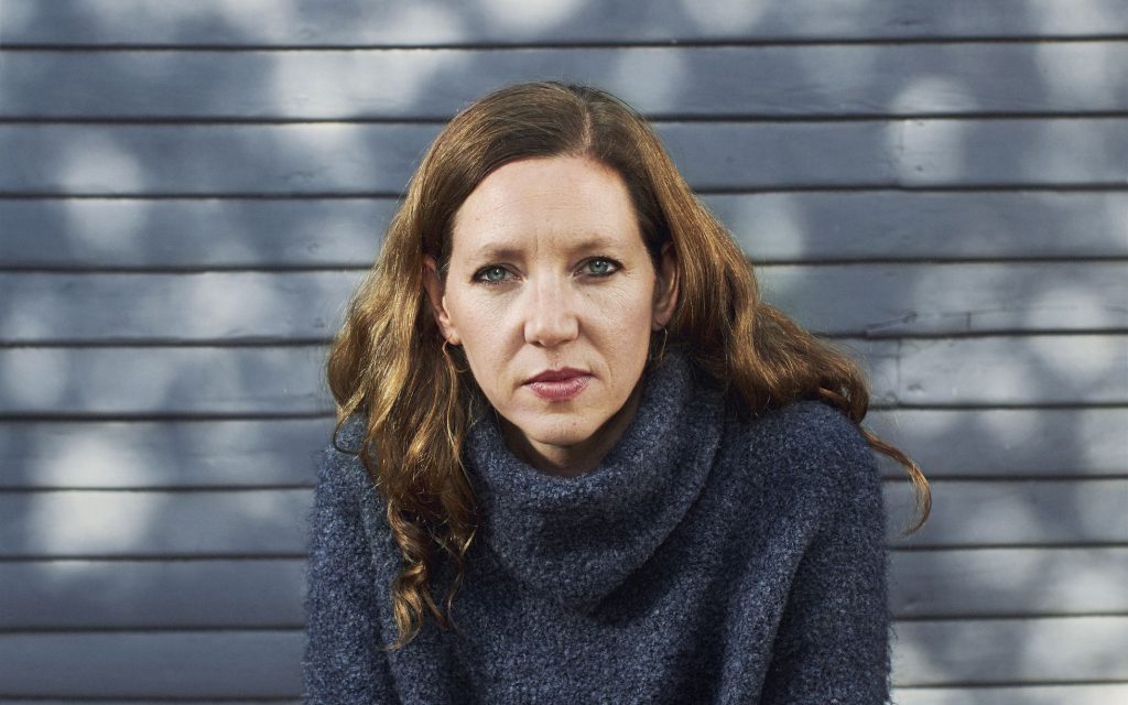 Maggie Nelson, crítica cultural y teórica, con cabello castaño ondulado y suéter gris oscuro frente a pared azul