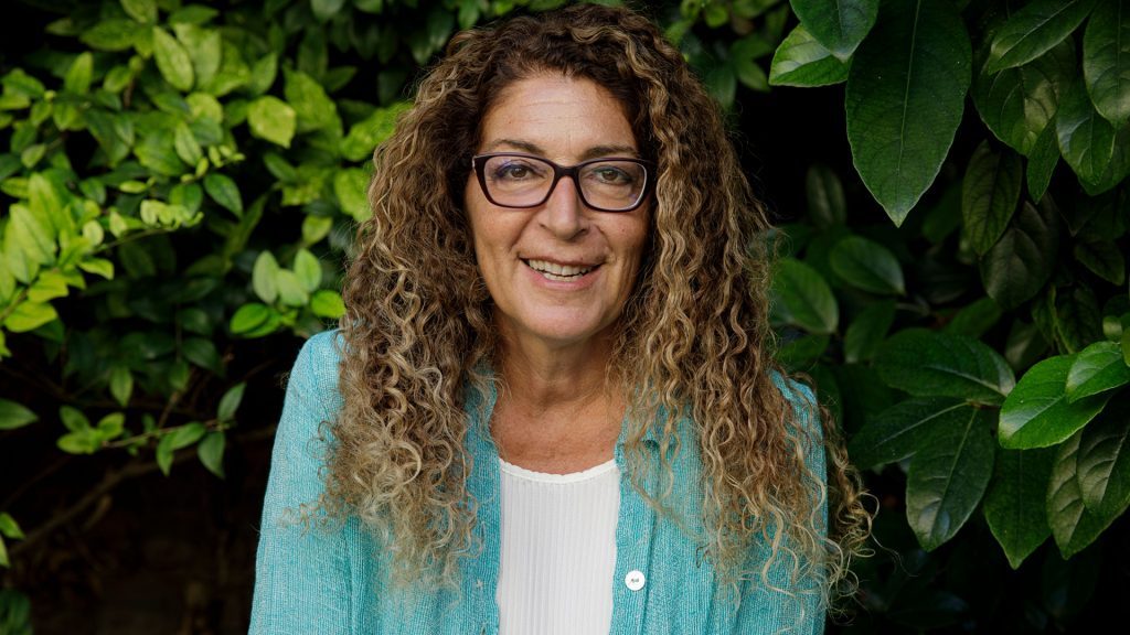 Melania G. Mazzucco, autora, con gafas y cardigan azul claro, frente a fondo de hojas verdes