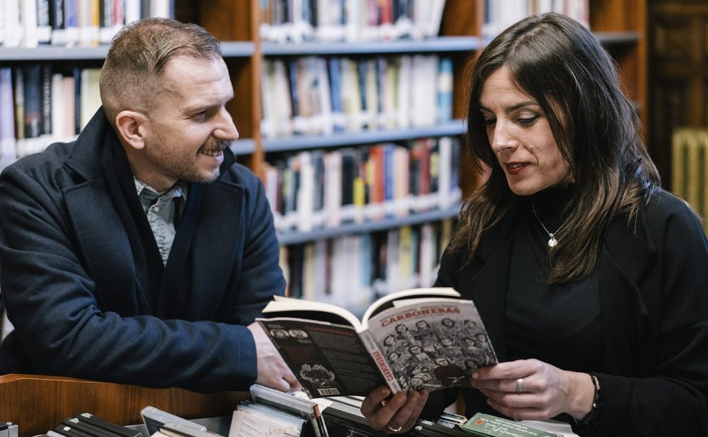 Dos personas leen juntas un libro en una biblioteca; ambiente académico con estanterías de libros de fondo