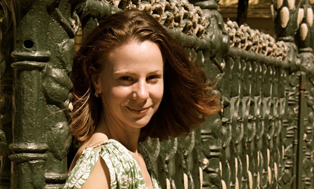 Mujer joven sonríe ante pared de piedra tallada con motivos decorativos, vistiendo blusa clara