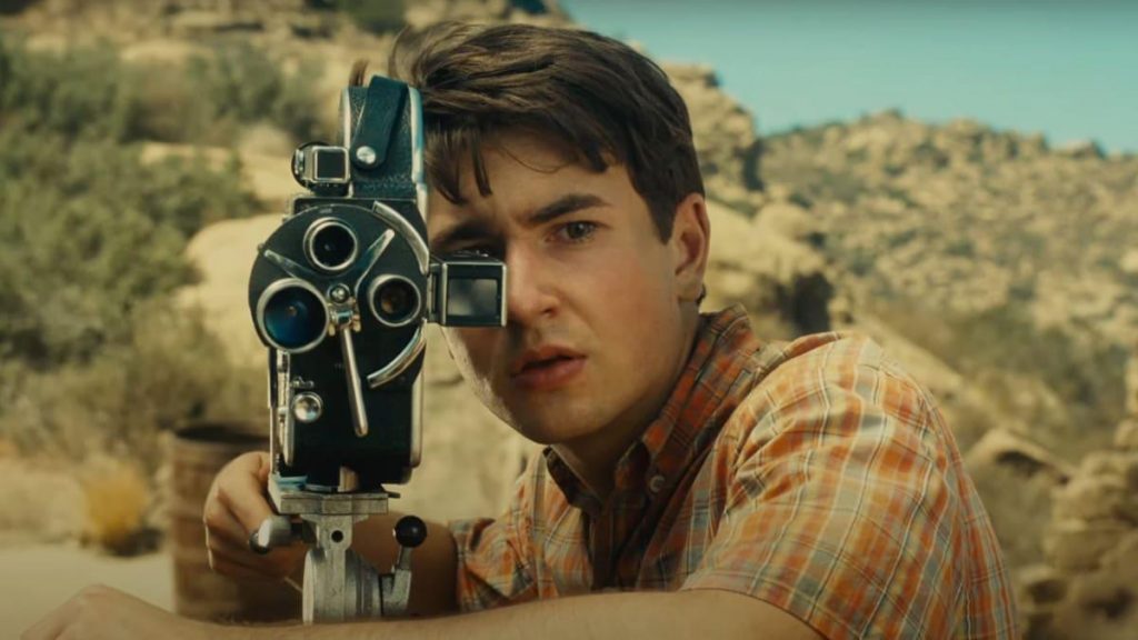 Joven sosteniendo una cámara de cine vintage en primer plano, con paisaje desértico de colinas al fondo