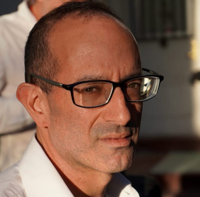 Hombre de mediana edad con gafas negras y camisa blanca, sonriendo levemente hacia la cámara en ambiente interior