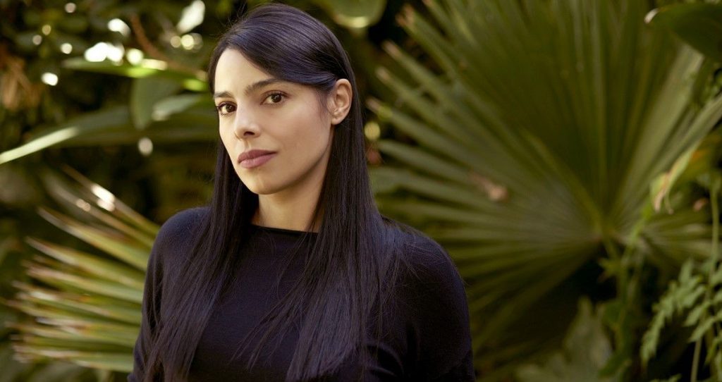 Margarita García Robayo en primer plano, con cabello largo negro, viste ropa oscura y está rodeada de vegetación