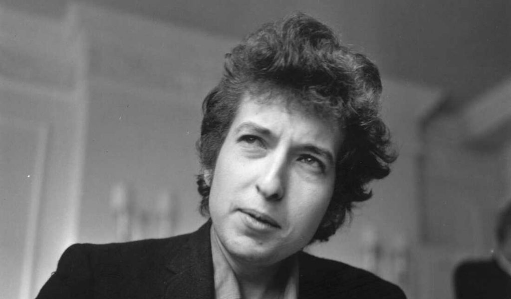 Bob Dylan en fotografía en blanco y negro, con cabello oscuro ondulado, mirando hacia un lado con expresión