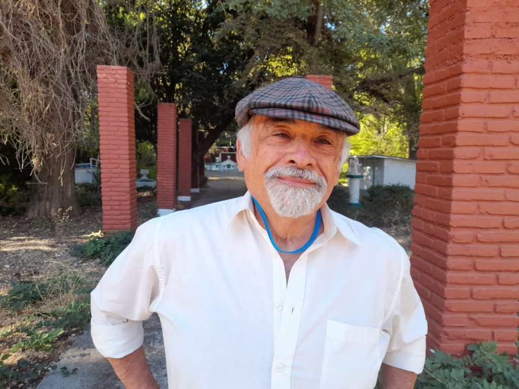 Juan Mihovilovich, hombre de mediana edad con gafas y camisa blanca, frente a pared de ladrillo rojo