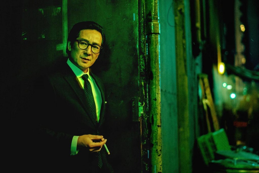 Hombre con gafas en traje oscuro junto a pared verde iluminada, con expresión seria en ambiente nocturno