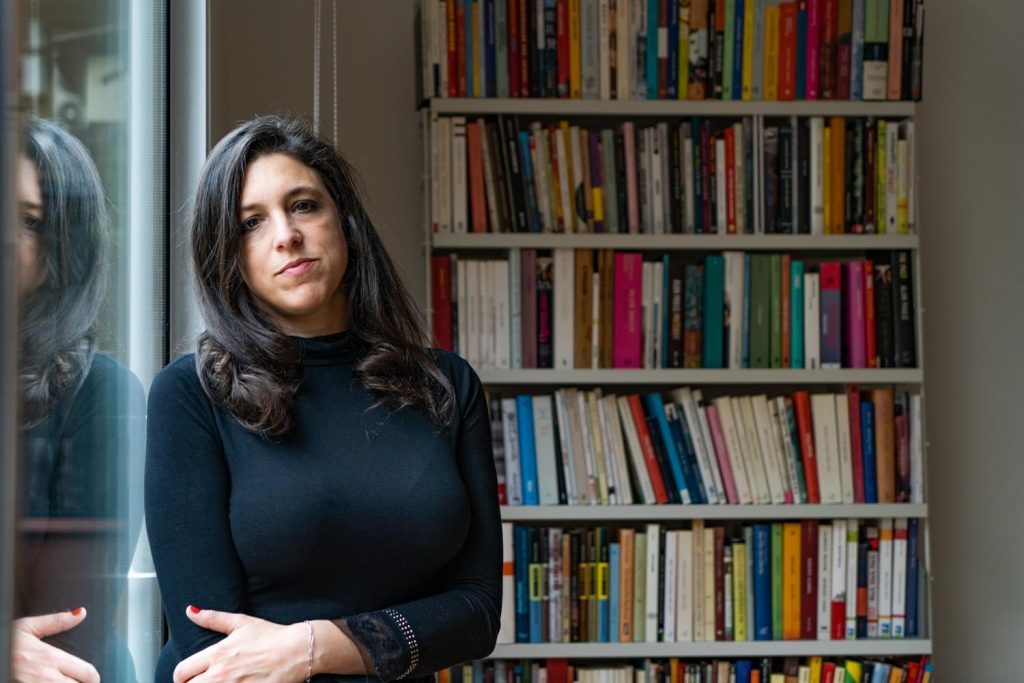 Ariana Harwicz junto a estantería de libros, mirando hacia la cámara en ambiente interior