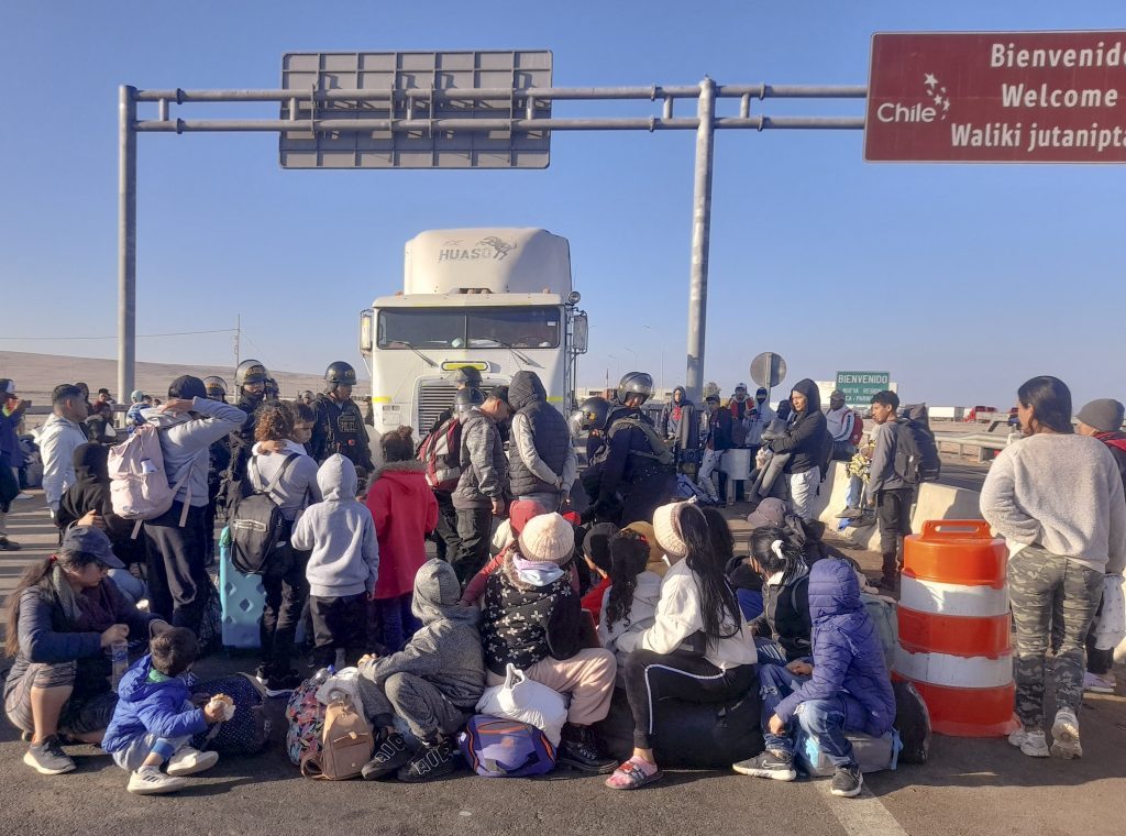 Grupo de migrantes reunidos bajo una carretera elevada, con maletas y pertenencias, esperando en condiciones precarias