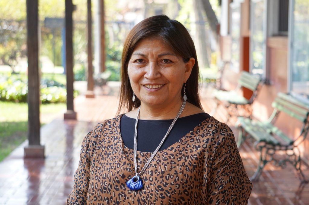 Mujer sonriente con largo cabello castaño, collar azul y blusa marrón, frente a arquitectura colonial de fondo
