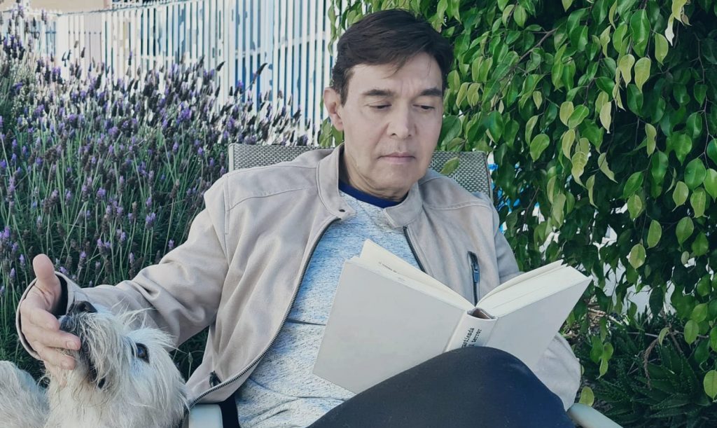 Jaime Vieyra sostiene un periódico mientras lee, sentado frente a una pared con plantas trepadoras verdes