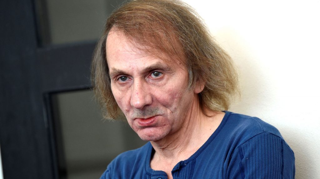 Hombre de cabello largo rojizo y barba, vistiendo camisa azul oscura, en primer plano contra fondo neutro