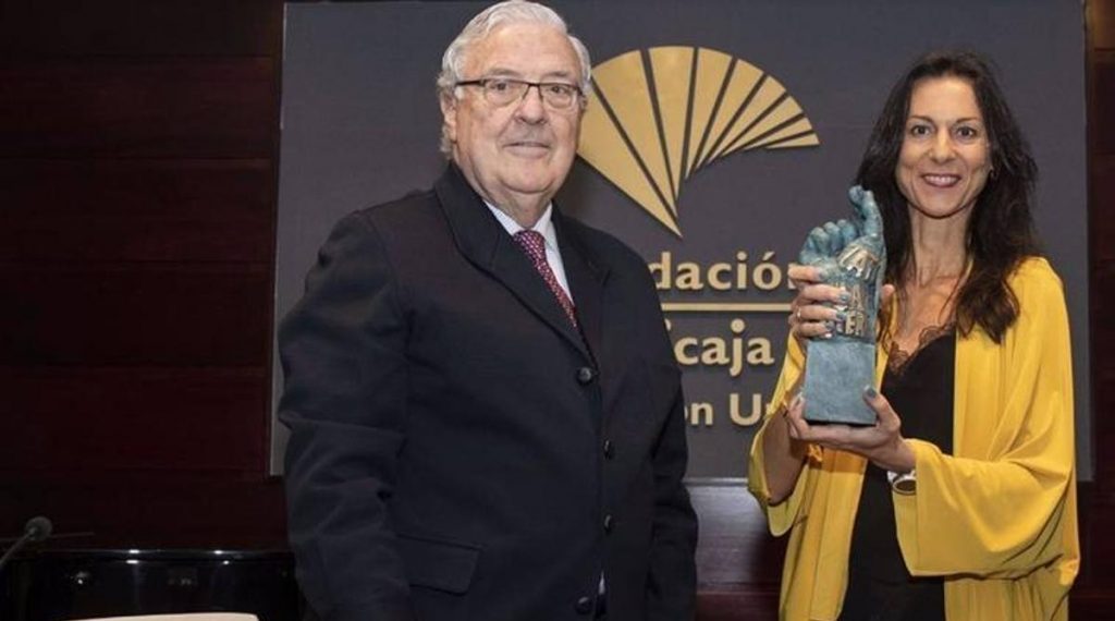 Hombre de anteojos con traje oscuro y mujer de blusa amarilla sostienen trofeo dorado en evento cultural