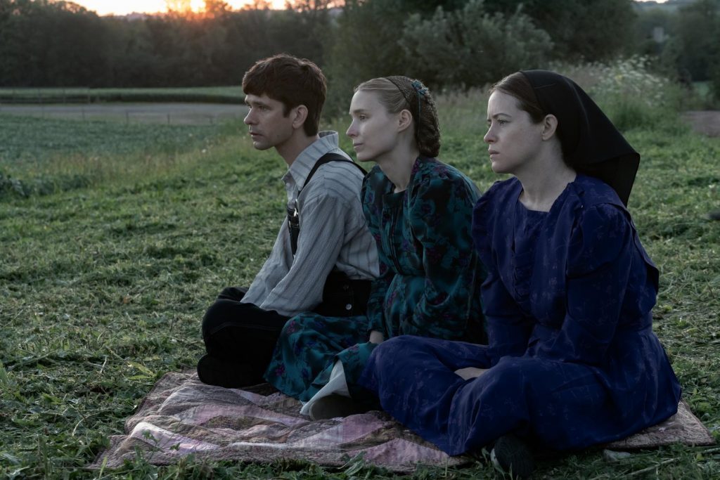 Tres jóvenes sentadas en un campo al atardecer, mirando hacia el horizonte con expresión seria y reflexiva