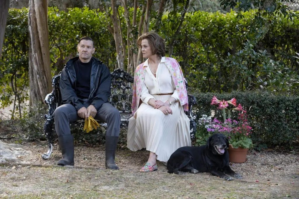 Hombre y mujer sentados en un banco de jardín junto a flores y arbustos, con un perro negro a sus pies