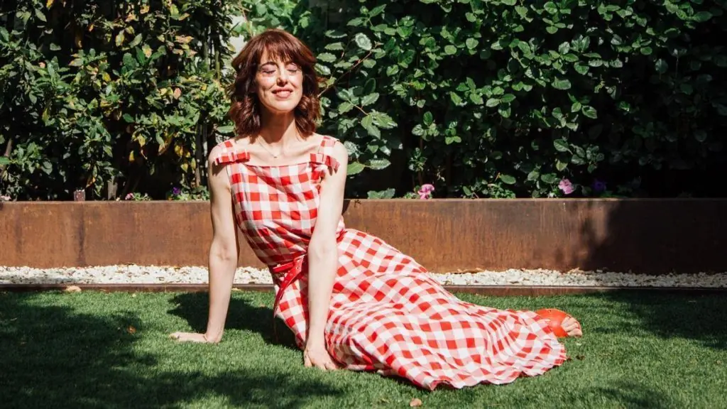 Irene Vallejo recostada en el césped con vestido blanco y rojo a cuadros, sonriendo hacia la cámara con follaje verde