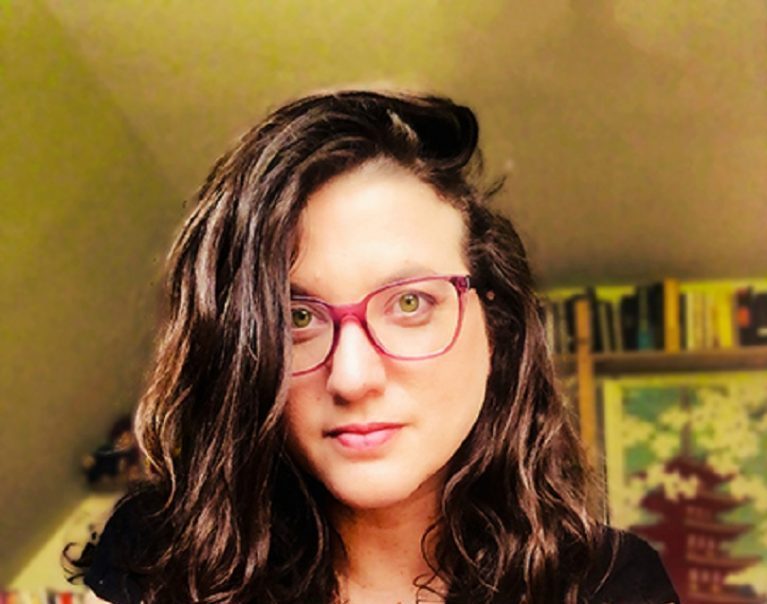 Mujer con lentes rosas y cabello negro largo frente a fondo amarillo con libros