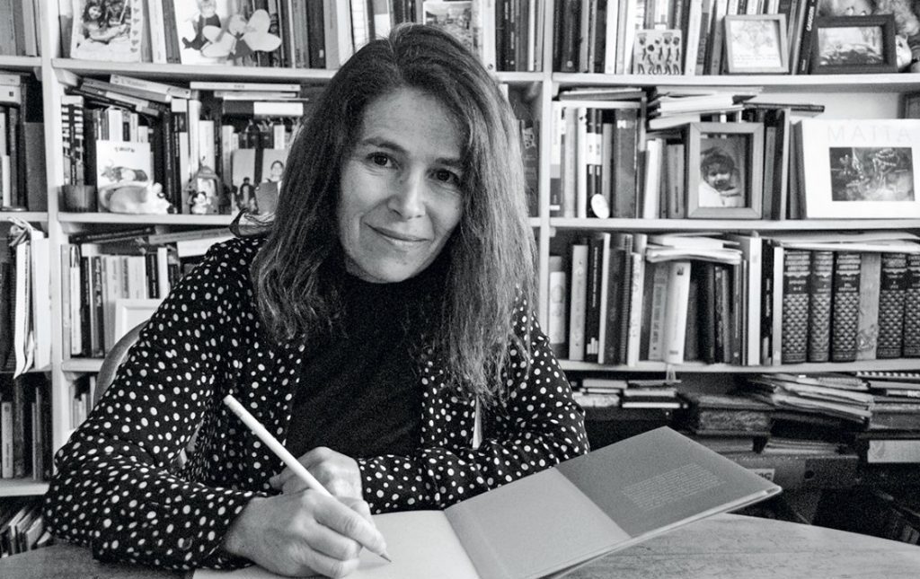 Marilú Ortiz de Zárate escribiendo en un libro ante estantería con libros de fondo, fotografía en blanco y negro