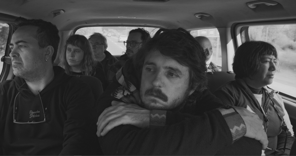 Cinco hombres en el interior de un automóvil en blanco y negro, con expresiones tensas y contemplativas