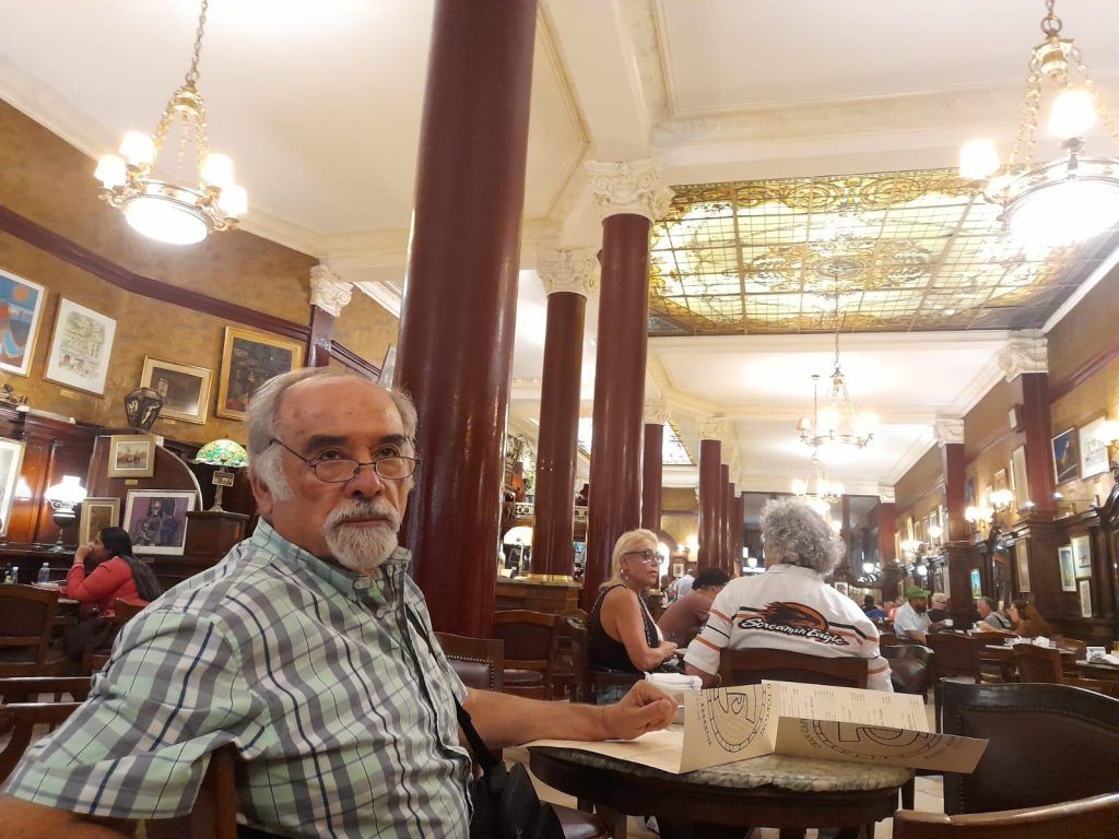 Juan Mihovilovich en interior de bar o cafetería con columnas de madera, mesas y otras personas al fondo