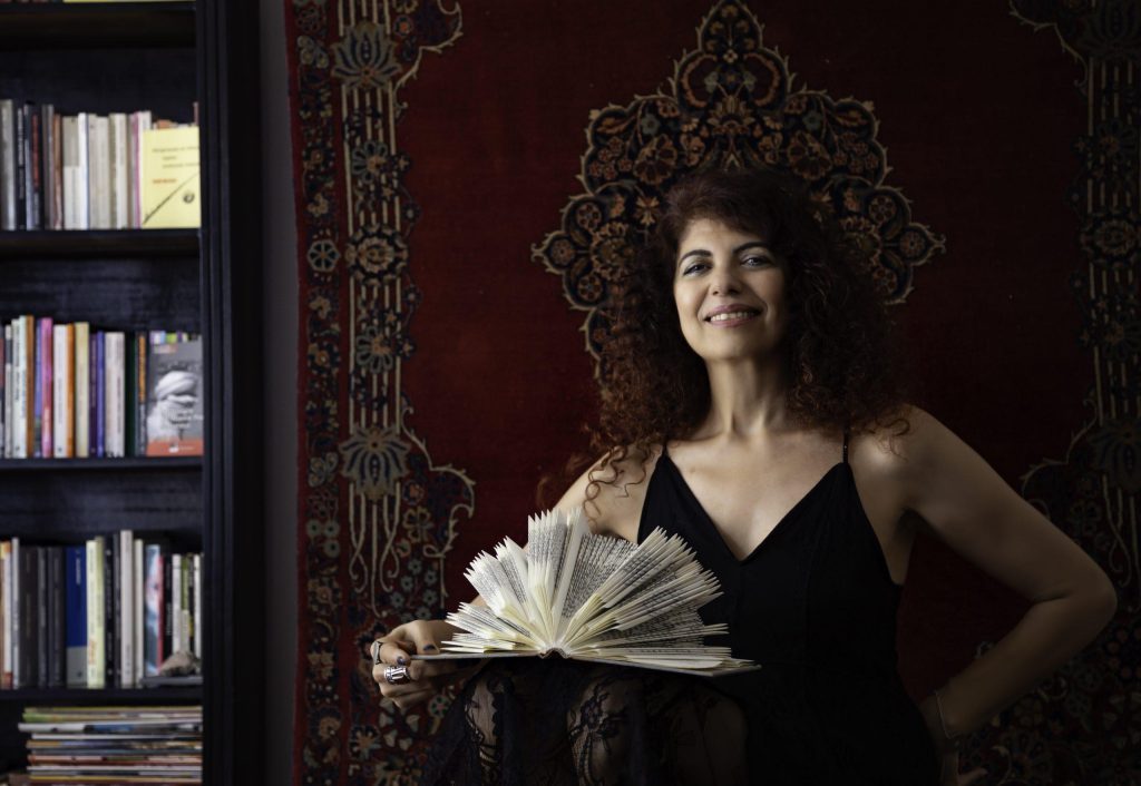 Mujer sosteniendo un libro abierto frente a fondo rojo decorado, con estantería de libros al fondo