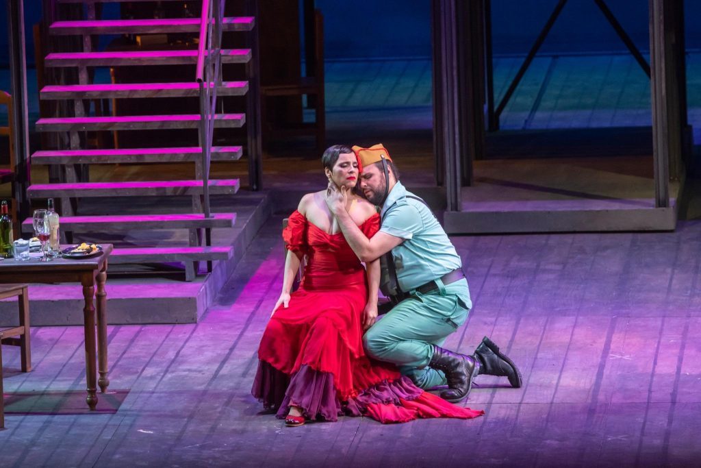 Escena de la ópera Carmen: mujer en vestido rojo y hombre en traje claro arrodillado frente a ella en escenario teatral
