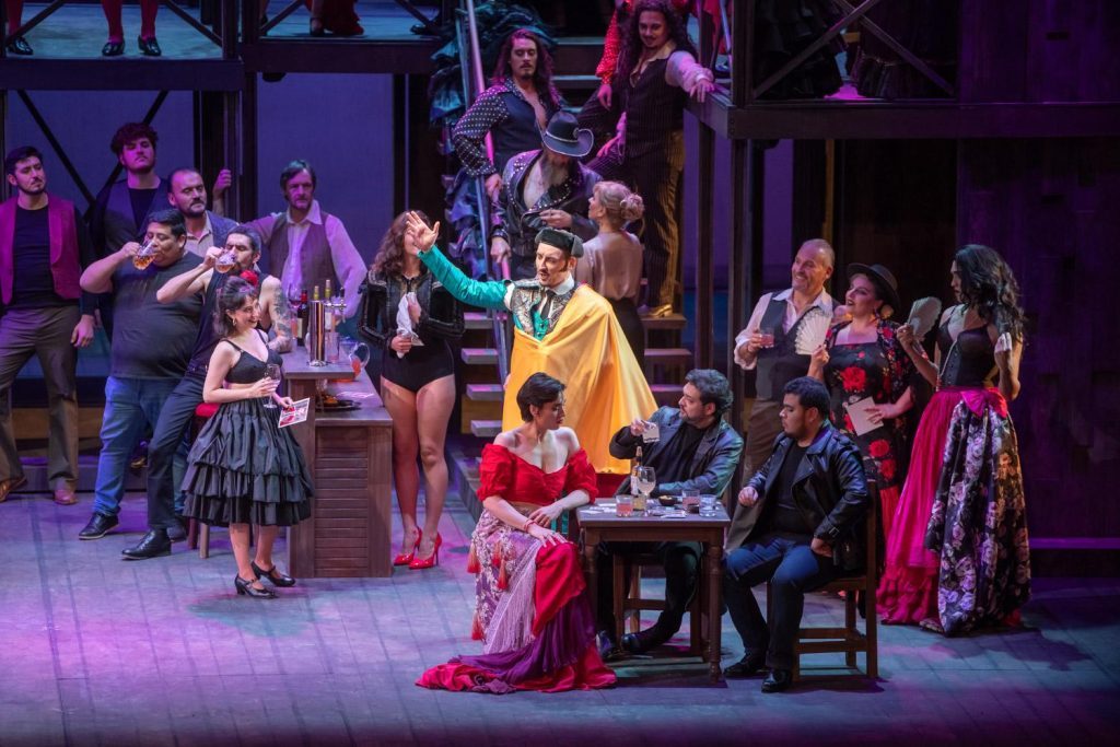 Escena teatral de Carmen con actores en escenario iluminado, algunos en primer plano con vestuario rojo y amarillo