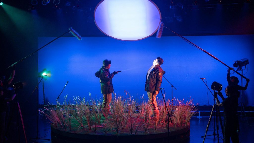 Dos actores en escena bajo iluminación azul con plantas rojas, cámaras de filmación y luces de estudio de fondo