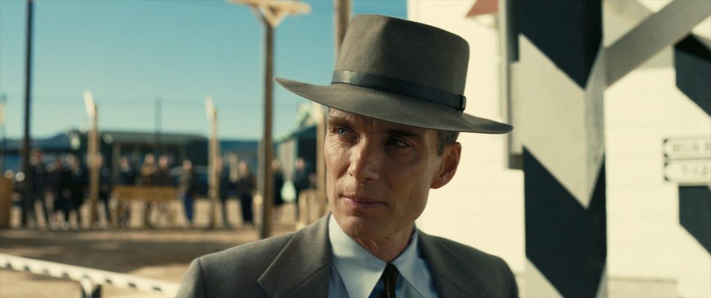 Hombre con sombrero y traje oscuro en escena de película, con edificios al fondo en ambiente desértico