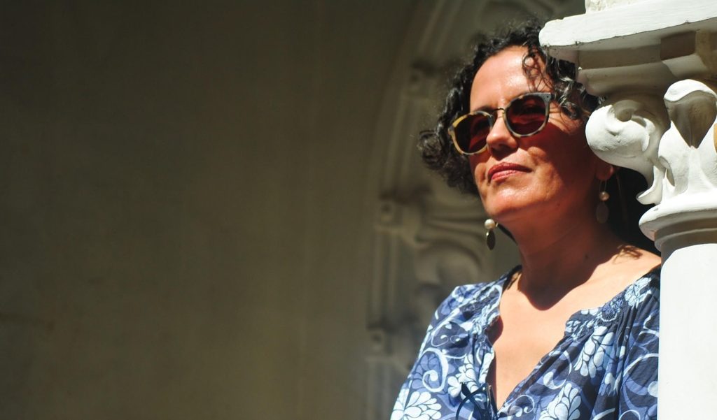 Mujer con gafas de sol rojas y blusa azul estampada, sonriendo junto a una pared blanca con objetos decorativos