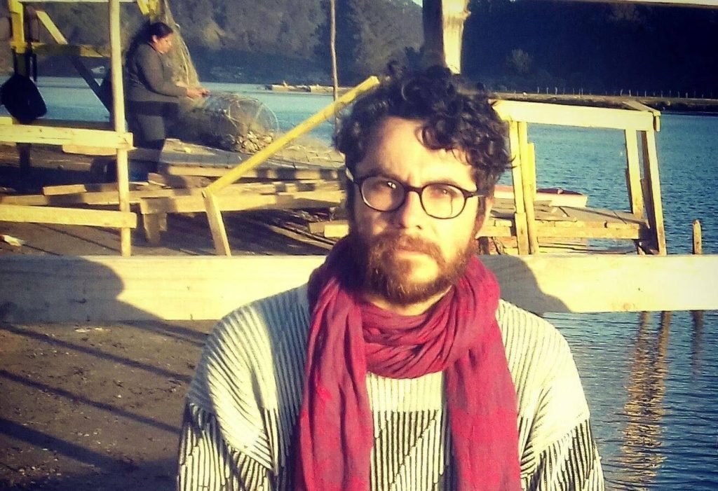 Eduardo Serrano con gafas, bufanda roja y camisa a rayas, frente a embarcaciones en agua