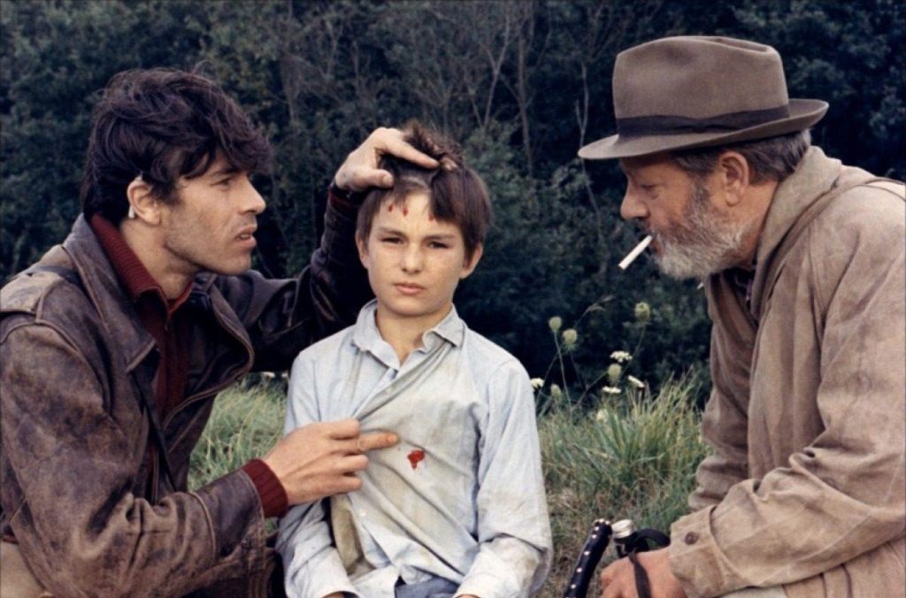 Tres hombres en una escena rural: dos adultos conversan con un niño entre árboles y vegetación natural