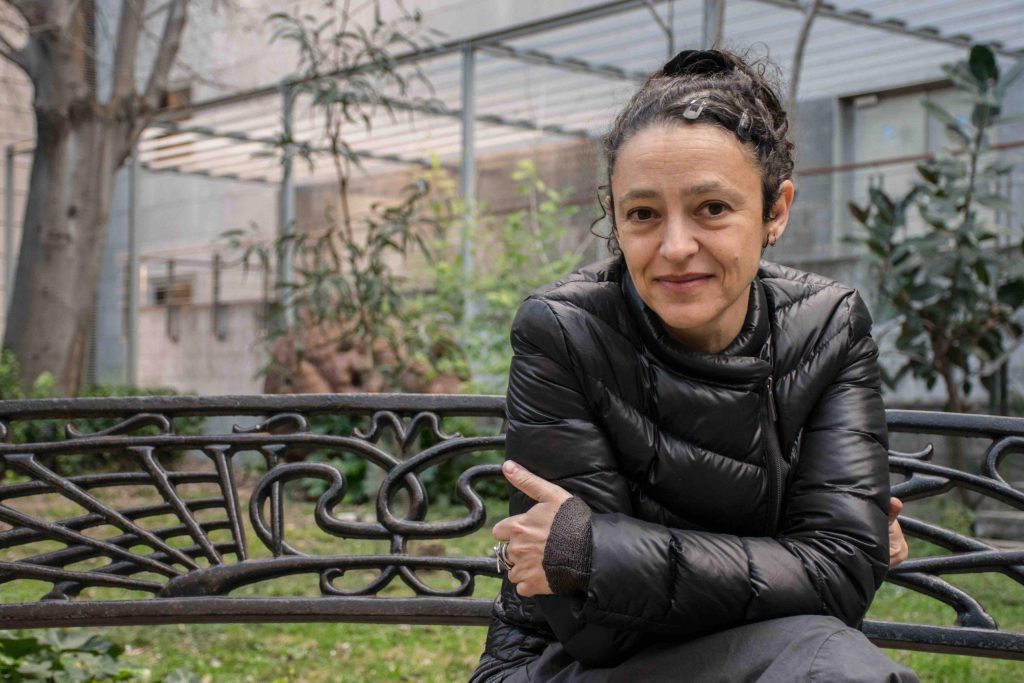 Lina Meruane apoyada en una barandilla de hierro forjado en un jardín, con invernadero de fondo