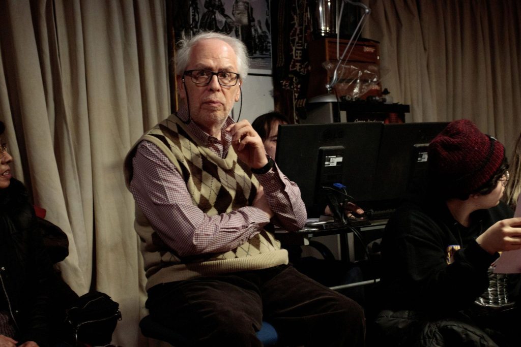 Manuel Rodríguez con gafas, camisa marrón y chaleco, sentado frente a cámara de cine en estudio