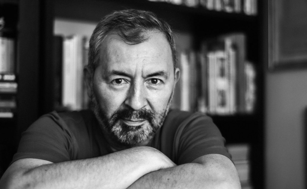 Marcelo Simonetti, escritor chileno, retrato en blanco y negro con brazos cruzados frente a estantería de libros