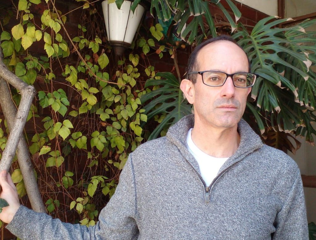 Nicolás Poblete Pardo con gafas y suéter gris ante pared con plantas verdes