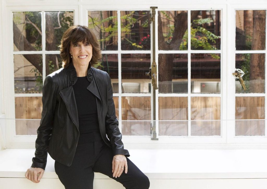 Nora Ephron apoyada en una ventana de marcos blancos, vistiendo chaqueta negra, sonriendo a la cámara