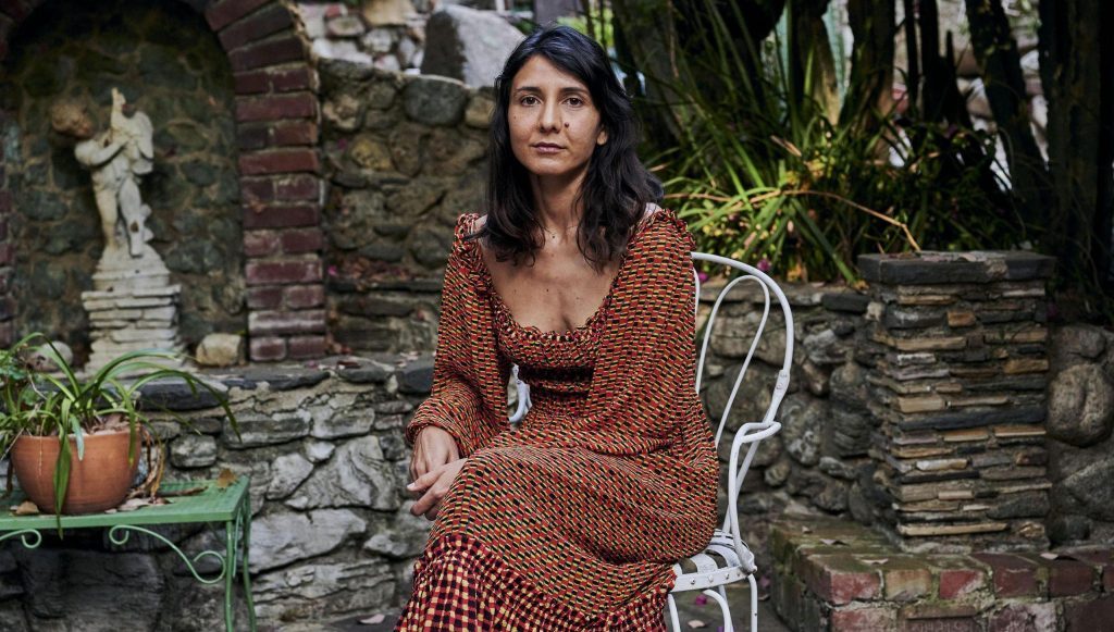 Ottessa Moshfegh sentada en una silla blanca en jardín con plantas y estatua de fondo, vistiendo vestido marrón tejido