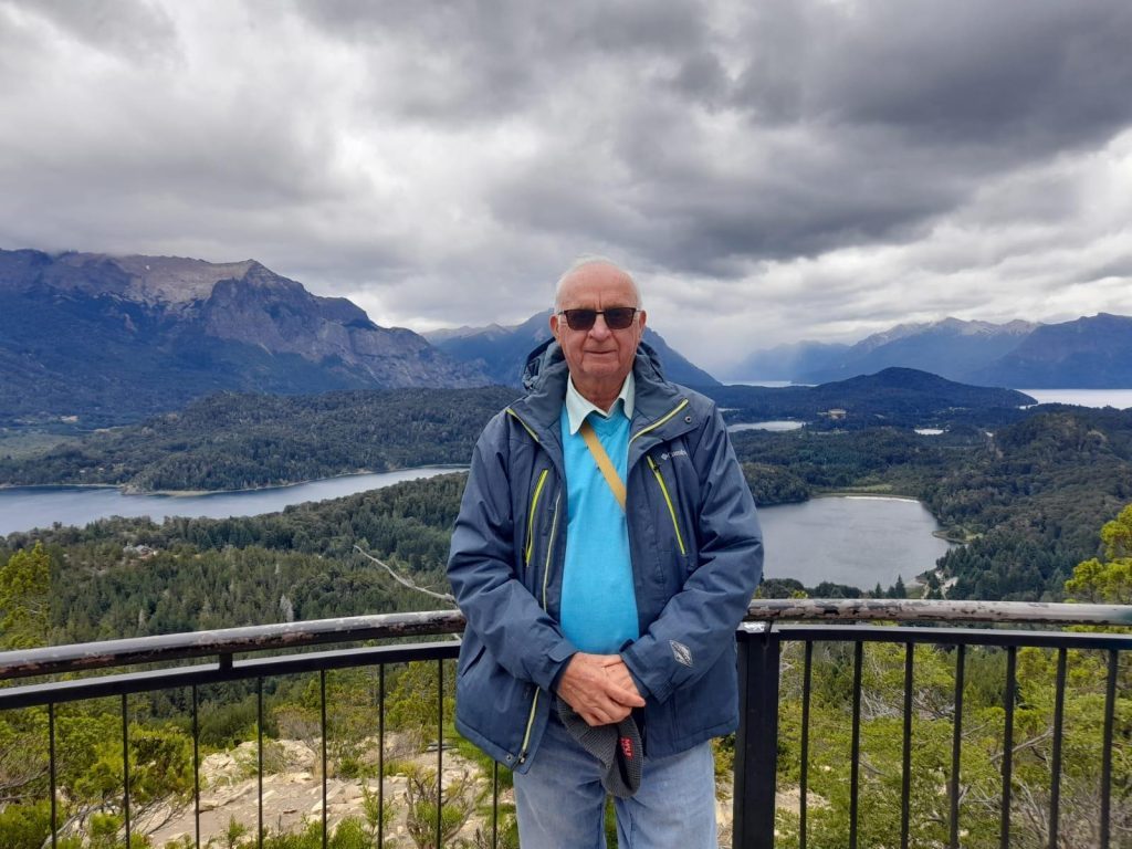 Rubén con gafas de sol y chaqueta azul en mirador de montaña, lakes y bosques al fondo bajo cielo nublado