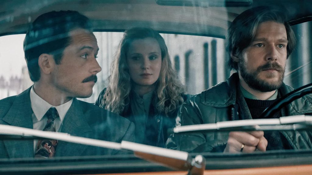 Tres personajes en el interior de un automóvil durante la Guerra Fría, observando hacia adelante con expresión seria