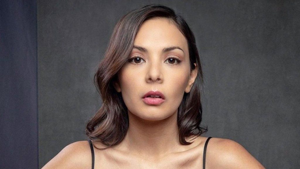 Retrato de Carolina Arredondo, mujer de cabello castaño ondulado, maquillaje en tonos púrpura, sobre fondo gris neutro