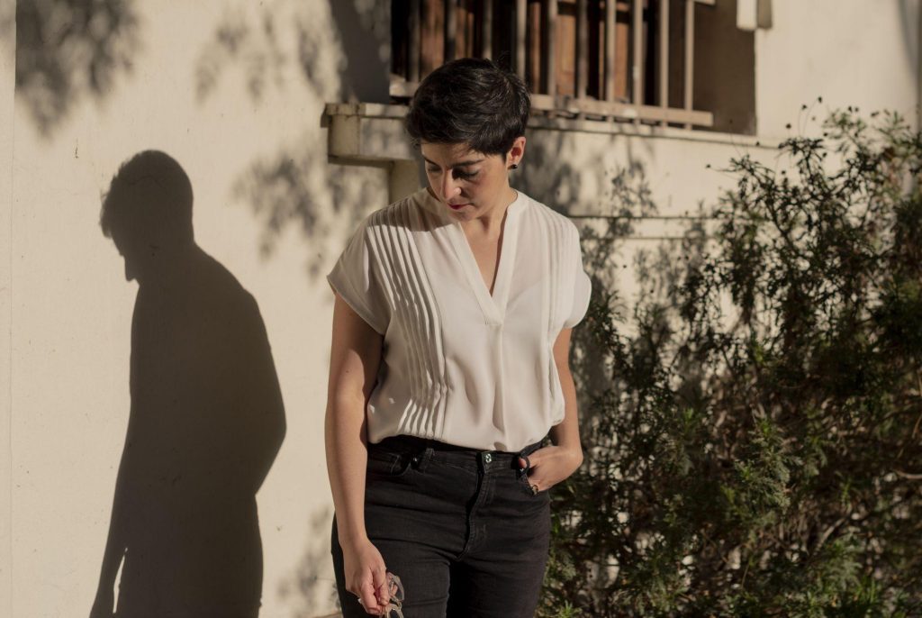 Alia Trabucco viste blusa blanca y pantalón negro, de pie ante pared blanca con sombra y plantas de fondo
