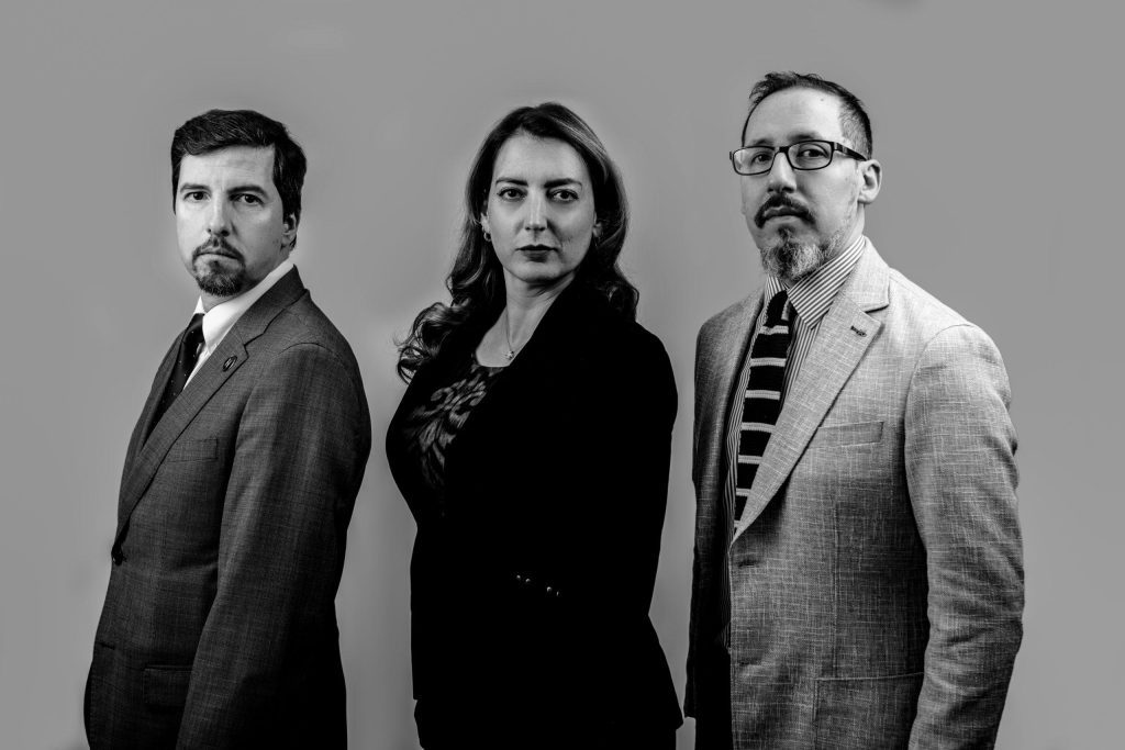 Tres personas en retrato formal en blanco y negro: hombre de bigote a la izquierda, mujer al centro y hombre con gafas
