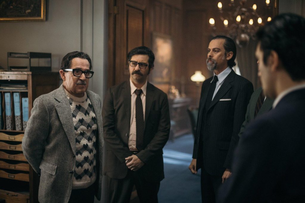 Cuatro hombres en traje conversan en un interior elegante con lámpara de araña, escena de la película Los mil días